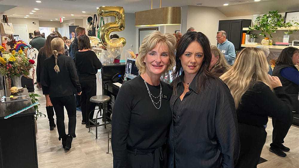 Two women posing at an event with a crowd and large gold number "5" balloon in the background.